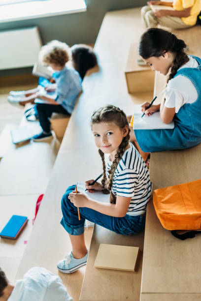 istock high angle view of schoolgirl with notebook studying on tribune at school corridor while her classmates sitting on background 1031956080