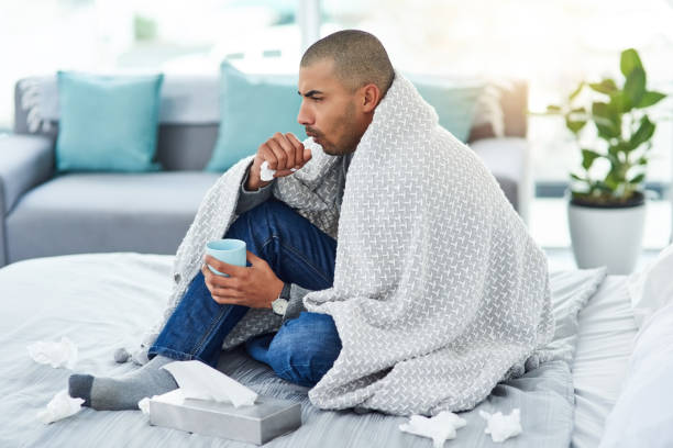 He's a little under the weather Full length shot of a young man sitting on his bed while feeling unwell at home bronchitis stock pictures, royalty-free photos & images