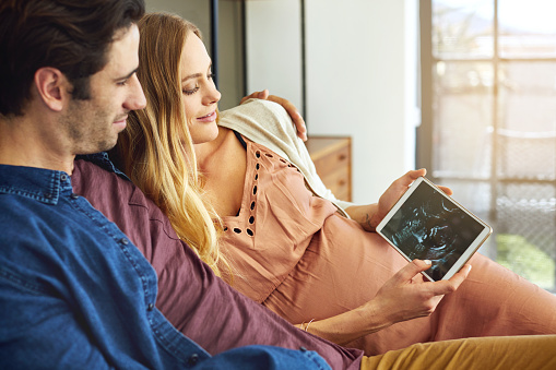 Shot of a young man and his pregnant wife looking at a sonogram on a digital tablet on the sofa