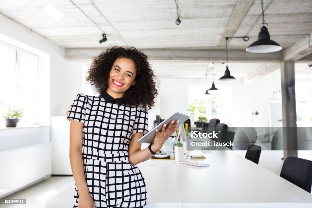 Female designer in modern work place Portrait of beautiful young woman with curly hair standing at a table with digital tablet in hand. Female designer in modern work place. 20-24 Years Stock Photo