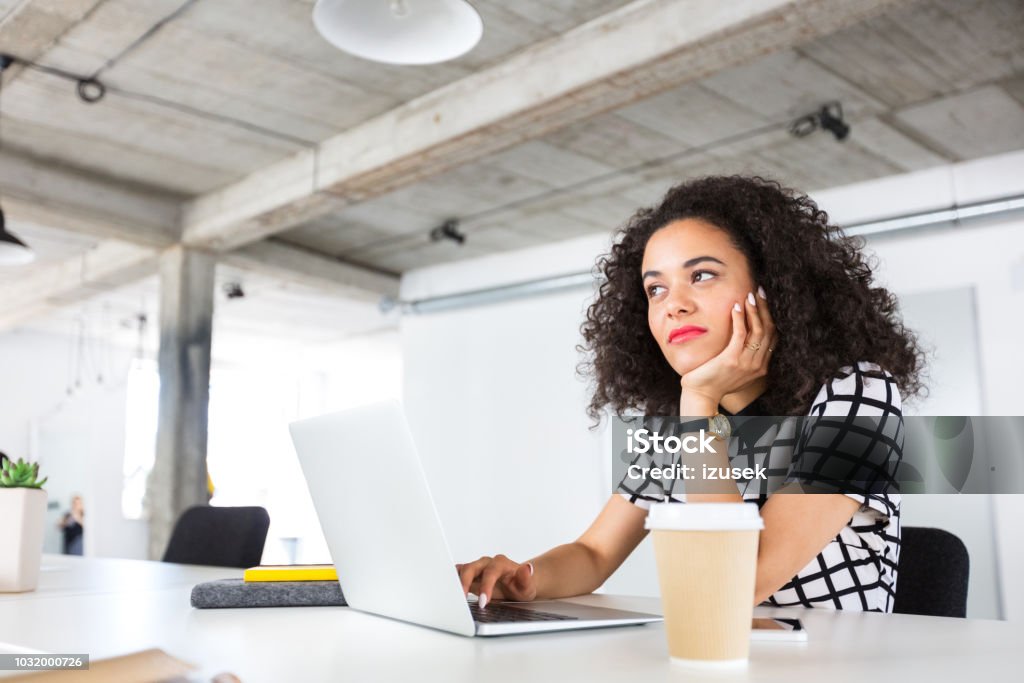 Creative professional with laptop looking away and thinking Portrait of attractive young female creative professional sitting at her desk with laptop looking away and thinking. Woman with curly hair working on laptop at office. Contemplation Stock Photo