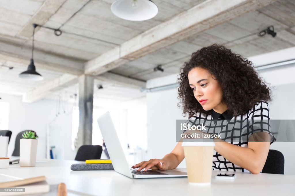 Woman designer working on laptop at office Portrait of beautiful young woman with curly hair sitting at her desk and working on laptop. Female designer working on laptop computer at office. 20-24 Years Stock Photo