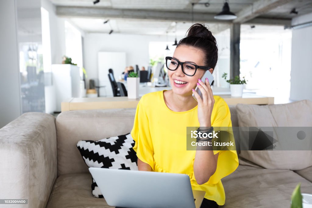 Businesswoman working in office Portrait of cheerful businesswoman sitting on couch in office working on laptop and talking on cell phone Women Stock Photo