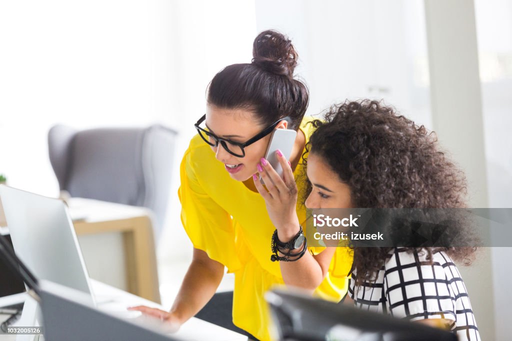 Young businesswomen working together in the office Two young businesswomen working together in the office Female working on computer with colleague talking on mobile phone. Customer Stock Photo