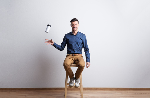 Portrait of a young handsome man in dark blue shirt sitting on a stool in a studio, trying to catch a travel coffee mug.