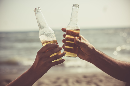 Close-up shot of male and female hand doing celebratory toast with beer bottles at the beach.