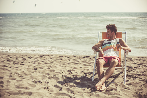 Copy space shot of a young man sitting on deck chair and drinking beer on the beach. Sea against sky in the background.