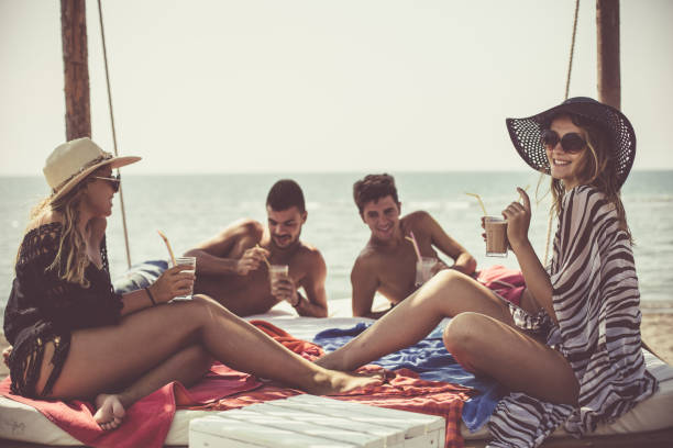 Your young friends are relaxing and drinking refreshing beverages on beach bed. Scenic view of sea against sky in the background.