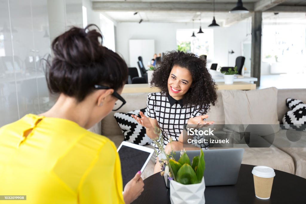 Design professionals discussing in office Two women working in office and discussing work. Smiling woman talking with female colleagues using digital tablet in creative workplace. Digital Tablet Stock Photo