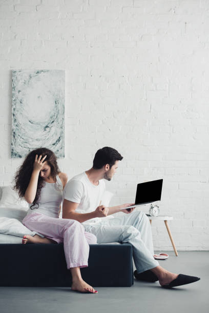 istock upset young woman sitting on bed while angry man using laptop with blank screen, relationship difficulties concept 1032026238
