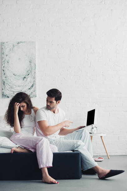 istock upset young woman sitting on bed while man pointing with finger at laptop with blank screen 1032026288