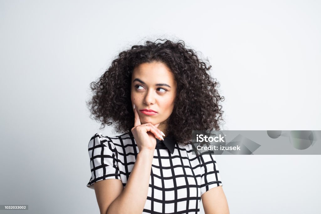 Thoughtful woman looking away on gray background Close up of young woman with hand on chin looking at copy space and thinking. Thoughtful female with curly hair on gray background. Asking Stock Photo
