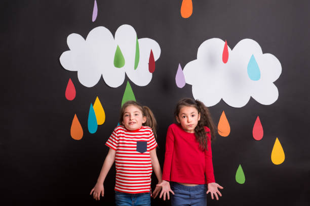 istock Small girls in studio, standing against black background with clouds and raindrops. 1032034030