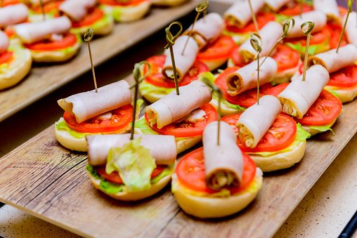 Close up image depicting a display of freshly prepared sandwiches, rolls and baguettes on display at a gourmet cafe. The sandwiches are loaded with a range of fillings including pastrami, smoked salmon, and mozzarella. The interior of the cafe, and a cafe worker, is defocused in the background.