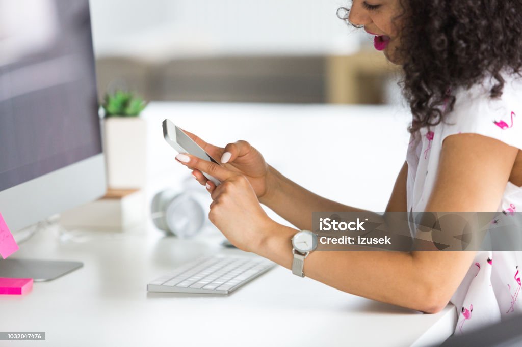 Female software engineer using mobile phone at her desk Young woman sitting at the desk using mobile phone. Female software engineer texting on her mobile phone. Close-up Stock Photo