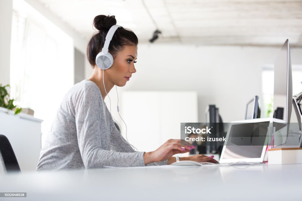 Application developer working on computer in office Female software developer sitting at office working on computer wearing headphones. Application developer working on a desktop computer in office. Computer Programmer Stock Photo
