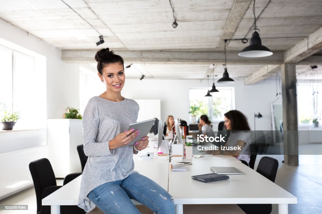 Beautiful young woman with digital tablet at office Portrait of beautiful young woman with digital tablet sitting at the table with coworkers working on computers in background. 20-24 Years Stock Photo