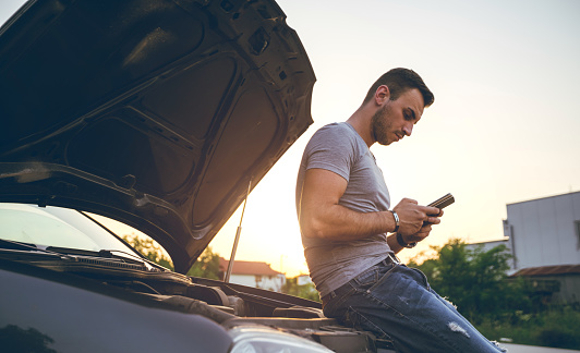 Young man is text messaging, standing next to a broken down car