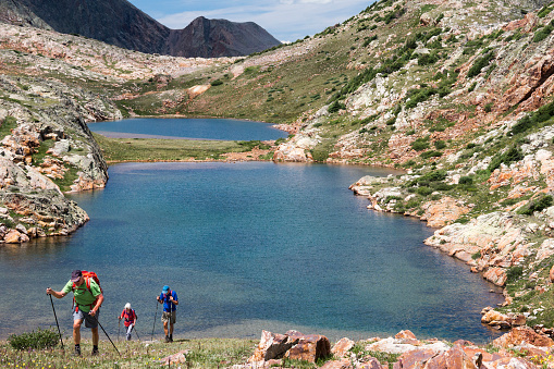 Three hikers in 60's use hiking poles to ascend a steep grassy slope from Eldorado Lake, at 12,500 ft elevation in the Grenadier Mountains, Continental Divide, San Juan Mountains, Rocky Mountains, Silverton, CO, USA