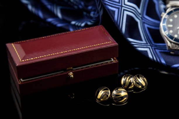 istock Golden cufflinks lying on a black reflective surface with a leather bound presentation box and a blue tie and stylish watch in background 1032089264
