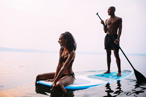 A young adult man and woman float on a standup paddleboard in the salt water of the Puget Sound, an inlet of the Pacific Ocean in Washington state, in the United States.  Rear view at sunset.  Shot at sunset.