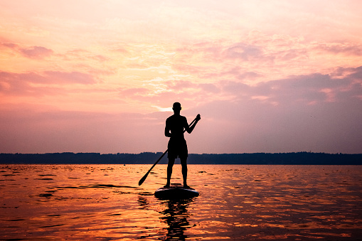 A young adult man floats on his standup paddleboard in the salt water of the Puget Sound, an inlet of the Pacific Ocean in Washington state, in the United States.  The sun casts pink and orange colors onto the water.