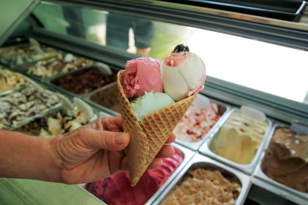 istock Female hand is holding a large strawberry, mint, vanilla with sour cherry ice cream in waffle cone. Ice cream fridge with steel service containers in background. Worker in ice cream shop. Pastry shop. 1032178708