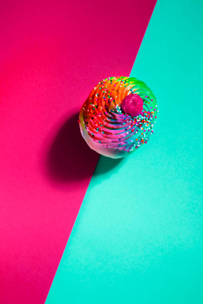 This is a photograph of a colorful sprinkled cupcake with a harsh shadow on a turquoise blue and red background and fits into the niche category of food pop art photography.