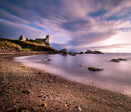 Long exposure of seascape featuring the old vintage ruins of Dunure Castle with wispy smooth clouds and ocean and sharp landscape of rocks and stony gravel.