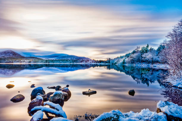 Photo of Beautiful winters day with soft clouds, snow on trees and rocks, reflections on calm water at Loch Morlich, Aviemore