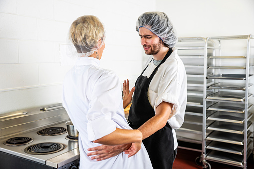 A male food service worker improperly touching a female colleague in a commercial kitchen.  He has his arm around her waist as she cooks at a stove. A concept on sexual harassment in the food service and restaurant industry.