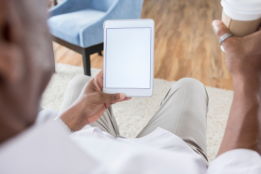 In this over the shoulder view, a man sits in a domestic looking room and holds a cup of coffee in one hand and a digital tablet in the other.  The screen is blank.