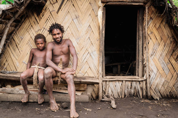 Tanna Island Tribe Young Father  and Son Kastom Village Vanuatu Tanna Island, Vanuatu - December 3rd, 2017: Smiling melanesian young man wearing tribal penis sheaths together with his son sitting on wooden bench in front of their Village Hut in remote Tribal Kastom Village. Tafea Province, Tanna Island, Vanuatu, Oceania. koteka stock pictures, royalty-free photos & images