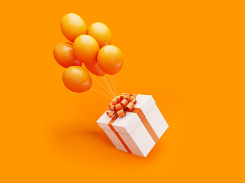 White gift box tied with orange colored ribbon is carried away by orange colored balloons on orange background.  Horizontal composition with copy space, Great use for Christmas and Valentine's Day related gift concepts.