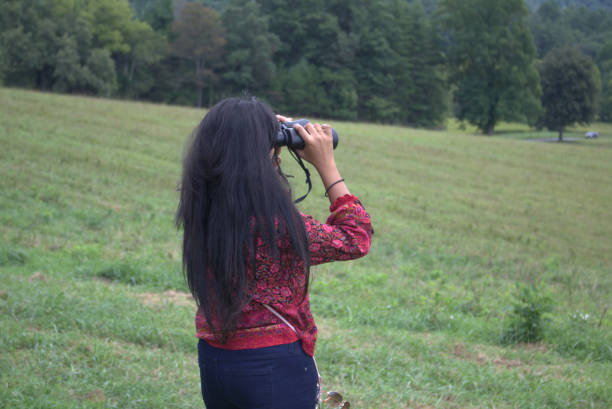 YOung woman with long, dark hair looking out over a field with binoculars stock photo