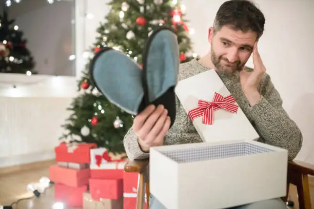 Caucasian man opening a Christmas present with disappointment.