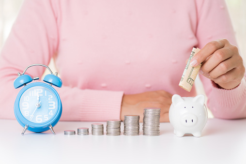 Woman hand putting America dollars banknotes money into piggy bank with stack of coins and alarm clock. Saving money wealth and financial concept.