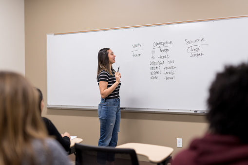 An ethnic female graduate teaching assistant stands at a large whiteboard and writes down Spanish verbs to conjugate for her students. The shot is over the shoulder of several students.