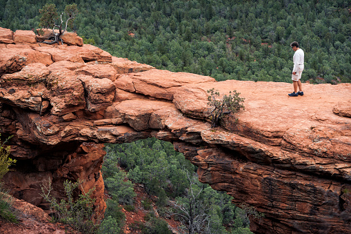 Apprehensive adult male cautiously and slowly walks out over the natural limestone archway known as Devil's Bridge while hiking on a spring day, Sedona, AZ, USA