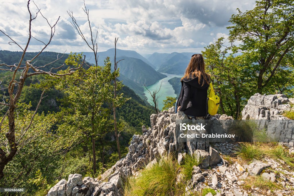 A tourist woman with a backpack sits on top of the mountain on Banjska Stena viewpoint A tourist woman with a backpack sits on top of the mountain on Banjska Stena viewpoint and looks ahead at the beautiful view in the distance, the mountains and the Drina River Hill Stock Photo