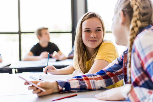 Female friends talking in classroom Female friends talking in classroom. Confident girls are sitting at table in high school. Students are wearing casuals. 15 year old blonde girl stock pictures, royalty-free photos & images