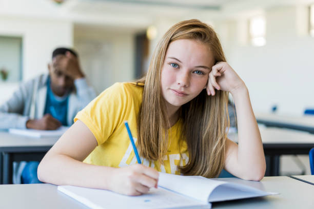 Confident female student writing in book at school Portrait of confident female student writing in book. Teenage girl is having long blond hair. She is sitting at desk in high school. 15 year old blonde girl stock pictures, royalty-free photos & images