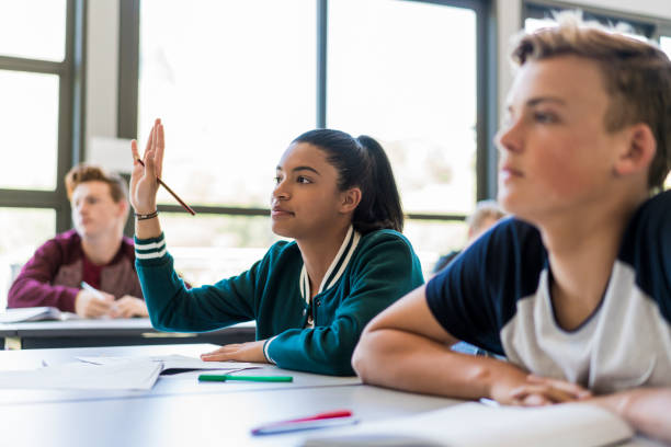 Female student raising hand to answer in classroom Female student raising hand to answer in classroom. Confident teenage girl is sitting with classmates. They are at desk in high school. hand raised classroom student high school student stock pictures, royalty-free photos & images