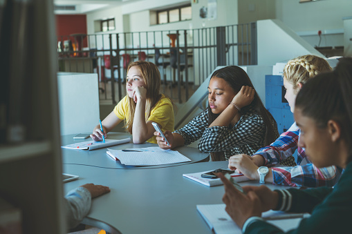 Teenage girls using phones while sitting with friends. Bored students are sitting at table. They are studying together in university.