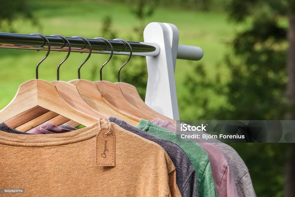 Organic clothes, t-shirts hanging on wooden hangers with green forest, nature in background. Organic clothes. Natural colored t-shirts hanging on wooden hangers in a row. Eco textile tag. Green forest, nature in background. Sustainable Resources Stock Photo