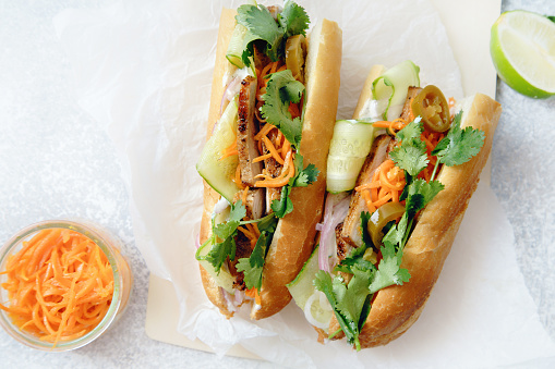 Classical banh-mi sandwich with sliced grilled pork tenderloin, shredded carrots and peeled cucumbers, jalapeno peppers and cilantro on white textured background. Top view, copy space