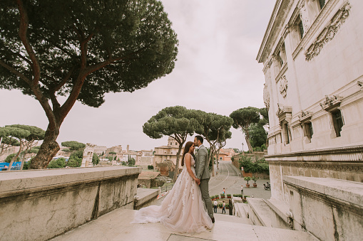 Young attractive newly married couple walking and posing in Rome with beautiful and ancient architecture on the background on their wedding day