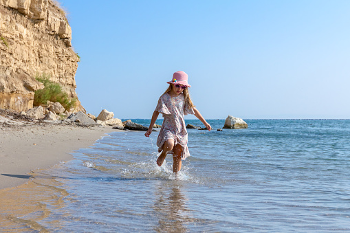 Child girl in summer dress with straw hat is running barefoot in shallow sea water