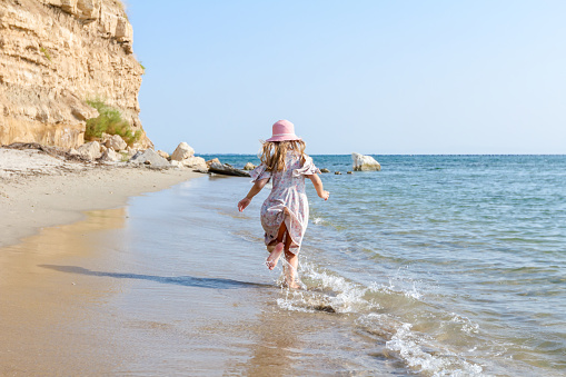 Child girl in summer dress with straw hat is running barefoot in shallow sea water