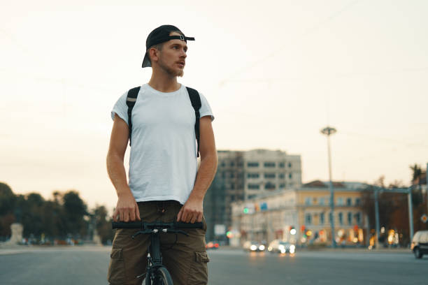Man riding bicycle in urban city holding hands on handlebar Portrait of a young man riding on bicycle in the city road, street with city far in the background. Male on black bicycle with white shirt, cap, backpack cycling to destination. Copy space on left. hipster bike stock pictures, royalty-free photos & images